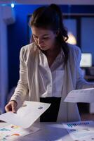 Stressed manager woman working with financial documents checking graphs, holding papers late at night. Business woman standing in start-up office doing overtime work to respect deadline. photo