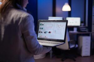 Back view of manager woman analysing financial statistics holding laptop standing in start-up business office late at night overworking. Tired focused employee using modern technology network wireless photo