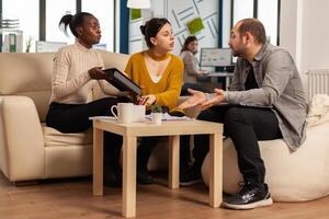 Diverse business people working in teamwork discussing project strategy using tablet sitting on couch at small table. Manager executive analysing financial data from black woman tablet at team meeting photo