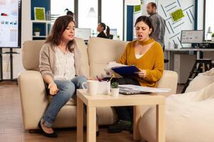 Woman leader yelling at employee sitting on couch in new start up business, upset of bad contract agreement. Women colleagues having big conflict, dispute about mistakes from project documents photo