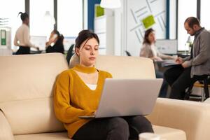 Concentrated business woman reading news on laptop, sitting on couch in busy start up company office, while diverse team analyzes statistics datas. Multiethnic team talking about project. photo