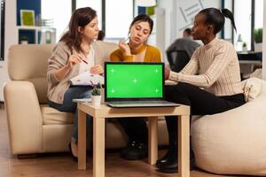 Laptop with chroma key display on table in startup business workplace. African manager discussing talking project statistics with employees on modren office sitting on couch. photo