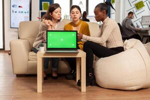 Laptop with green screen on table in start up company with multiethnic diverse coworkers working together behind it. African american woman discussing project strategy with colleques looking at tablet pc. photo