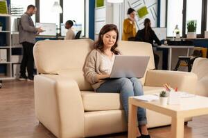 Business woman typing on laptop sitting on couch in start up office while diverse team working in background analysing statistics data. Multiethnic coworkers talking about startup financial company photo
