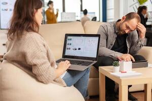 Back view of young business woman sitting on couch in office using laptop computer with graphics while colleagues working on background. Multiethnic coworkers planning new financial project photo