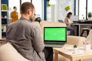 Over the sholder of manager sitting on couch holding laptop with green screen, mockup desktop while diverse team working on background. Multiethnic people planning project on chroma key display photo