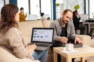 Back view of woman entrepreneur sitting on couch in office using laptop computer while colleagues working on background. Multiethnic coworkers planning new financial project in modern company photo