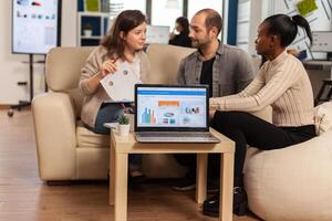 African woman talking about project, discussing start up ideas using laptop with graphics, diverse employees gathered in co-working process in company. Teamworkers helping each other finding strategy photo