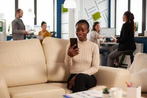 African manager woman discussing with remote colleagues on call holding smartphone using headphones sitting on couch in business modern office. Diverse coworkers planning new financial project photo