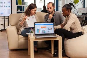 Diverse colleagues employees cooperating in office discussing at workplace, analysing graphs sitting on couch. Business coworkers planning start up business project, brainstorming in corporate office photo