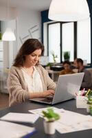 Satisfied manager checking financial statistics and graphs typing on laptop sitting at desk working in business start up company. Diverse team analyzes statistics data in modern office photo