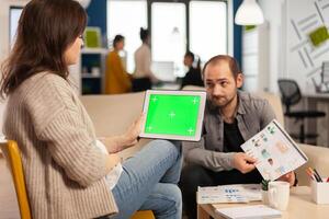 Business woman sitting on couch analysing financial statistics, holding tablet with green screen while diverse team working on background. Multiethnic coworkers planning project on chroma key display photo