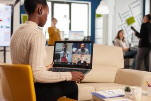 African manager woman discussing with remote colleagues using call holding laptop sitting on couch in business modern office. Diverse coworkers planning new financial project in modern company photo