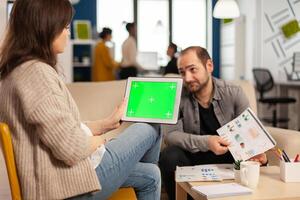 Over the sholder of manager woman sitting on couch talking with coworker holding tablet with green screen while diverse team working. Multiethnic people planning project on chroma key display photo