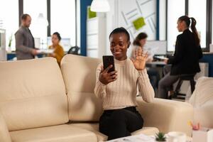 Black woman waving at camera explaining financial reports to remote manager on call holding smartphone using headphones sitting on couch. Diverse coworkers planning financial project photo