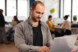 Enthusiastic business man writing a report sitting at table in modern startup business office sending mails typing on laptop, greeting colleague. Man working in financial company with diverse team. photo