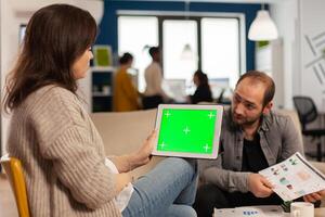 Back view of businesswoman sitting on couch using tablet with green screen talking with colleague while team working on background. Multiethnic coworkers planning project on chroma key display photo