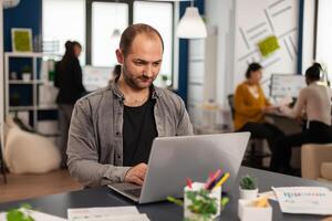 Man authentic manager sitting at table in modern startup business office sending mails typing on laptop. Business employee working in financial company with diverse team photo