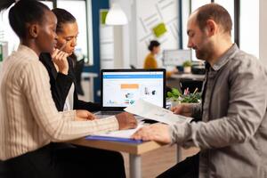 Busy multicultural, diverse employees analysing annual financial statistics sitting at desk in front of laptop holding documents searching business solutions. Team of businesspeople working in company photo