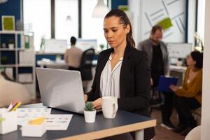 Concentrated business lady writing on laptop computer financial reports sitting at desk in busy start up office enjoying work in creative workplace. Diverse team analyzes statistics data in company photo