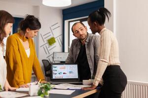 Diverse businesspeople communicating in start up business office standing over desk looking through financial documents. Multiethnic team analysing company informations in modern workplace photo