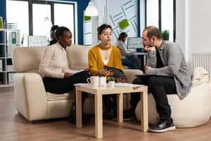 Concentrated multiethnic businesspeople reading annual financial report from tablet sitting on couch in modern startup business office. Diverse team of multiethnic businesspeople working in company photo