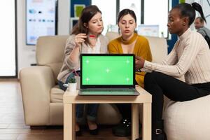 Laptop with green screen on table in start up company with multiethnic diverse coworkers working together behind it. African american woman discussing project strategy with colleques looking at tablet pc. photo