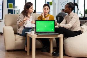 Laptop with chroma key display on table in startup business workplace. African manager discussing talking project statistics with employees on modren office sitting on couch. photo