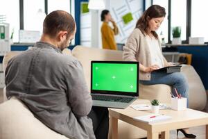 Over the sholder of manager sitting on couch holding laptop with green screen, mockup desktop while diverse team working on background. Multiethnic people planning project on chroma key display photo