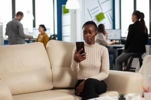 African manager woman discussing with remote colleagues on call holding smartphone using headphones sitting on couch in business modern office. Diverse coworkers planning new financial project photo