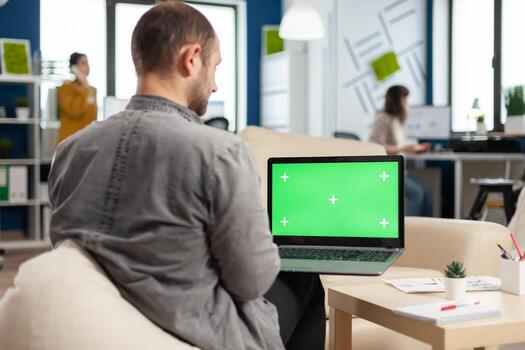 Business team sitting on couch analysing financial statistics, holding laptop with green screen while diverse team working on background. Multiethnic coworkers planning project on chroma key display photo