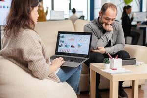Back view of woman entrepreneur sitting on couch in office using laptop computer while colleagues working on background. Multiethnic coworkers planning new financial project in modern company photo