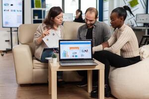 Diverse colleagues employees cooperating in office discussing at workplace, analysing graphs sitting on couch. Business coworkers planning start up business project, brainstorming in corporate office photo