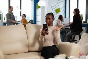 Black woman waving at camera explaining financial reports to remote manager on call holding smartphone using headphones sitting on couch. Diverse coworkers planning financial project photo