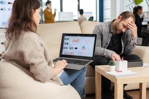 Back view of young business woman sitting on couch in office using laptop computer with graphics while colleagues working on background. Multiethnic coworkers planning new financial project photo