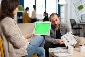 Business woman sitting on couch analysing financial statistics, holding tablet with green screen while diverse team working on background. Multiethnic coworkers planning project on chroma key display photo
