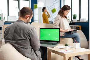 Back view of business man sitting on couch using laptop with green screen talking with colleague while team working on background. Multiethnic coworkers planning project on chroma key display photo