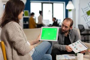 Over the sholder of manager woman sitting on couch talking with coworker holding tablet with green screen while diverse team working. Multiethnic people planning project on chroma key display photo
