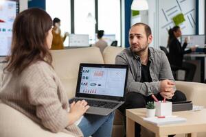 Back view of woman entrepreneur sitting on couch in office using laptop computer while colleagues working on background. Multiethnic coworkers planning new financial project in modern company photo