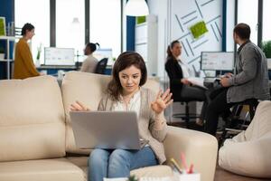 Manager woman sitting on couch holding laptop and talking on call during virtual conference working in business modern office. Diverse coworkers planning new financial project in modern company photo