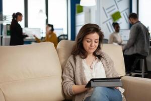 Employee taking break sitting on confortable couch holding tablet browsing, reading great news and smiling. Multiethnic creative coworkers planning new financial project working in open plan office photo
