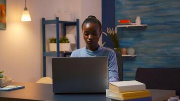 African woman coming in living room and typing on computer working from home overtime. Black entrepreneur sitting in personal workplace writing on keyboard looking at desktop respecting deadline. photo