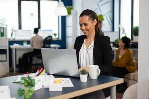 Concentrated business lady writing on laptop computer financial reports sitting at desk in busy start up office enjoying work in creative workplace. Diverse team analyzes statistics data in company photo