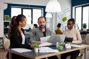 Happy diverse businesspeople analysing great annual financial reports sitting at desk using laptop. Man leader explainig project strategy using graphics, discussing project ideas, brainstorming photo