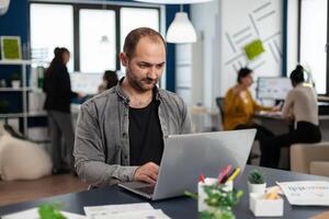 Man authentic manager sitting at table in modern startup business office sending mails typing on laptop. Business employee working in financial company with diverse team photo