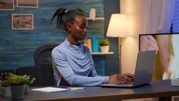 Black woman typing on computer and writing list of tasks on sticky notes putting it on laptop sitting on desk in living room workplace late at night. African freelancer working from home overtime photo