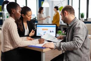 Busy multicultural, diverse employees analysing annual financial statistics sitting at desk in front of laptop holding documents searching business solutions. Team of businesspeople working in company photo