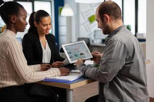 Enthusiastic diverse businesspeople reading annual financial report sitting at table in modern startup business office holding tablet and smiling. Team of multiethnic businesspeople working in company photo
