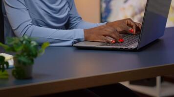 Close up of african woman typing on laptop sitting in living room late at night and working. Busy focused employee using modern technology network wireless doing overtime for job writing, searching photo