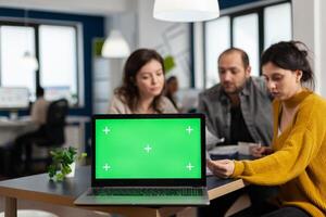Laptop with green screen chroma key display. mock up desktop ready for presentation placed on desk while business people work in background. Group of employees using notebook with isolated monitor photo